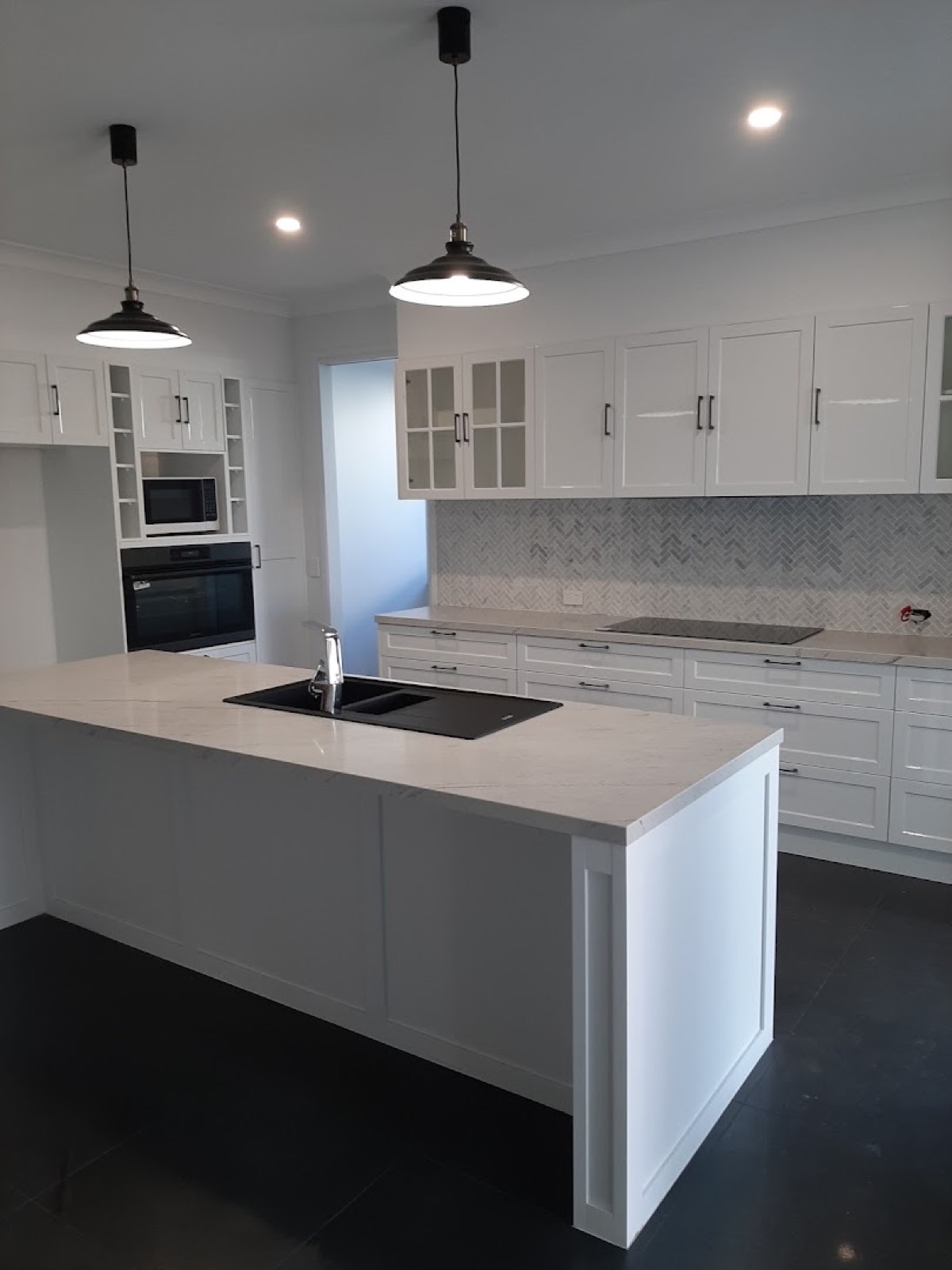 A white kitchen with a large island bench, glass wall cabinets and pendant lights